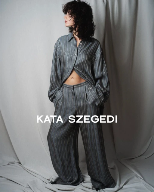A woman with curly dark hair poses in a grey striped blouse and pants set in a studio with neutral-toned draped backdrop and soft lighting.