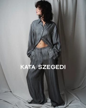 A woman with curly dark hair poses in a grey striped blouse and pants set in a studio with neutral-toned draped backdrop and soft lighting.