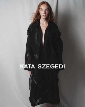 Model with red hair wearing a black sheer textured coat standing against a pale fabric backdrop