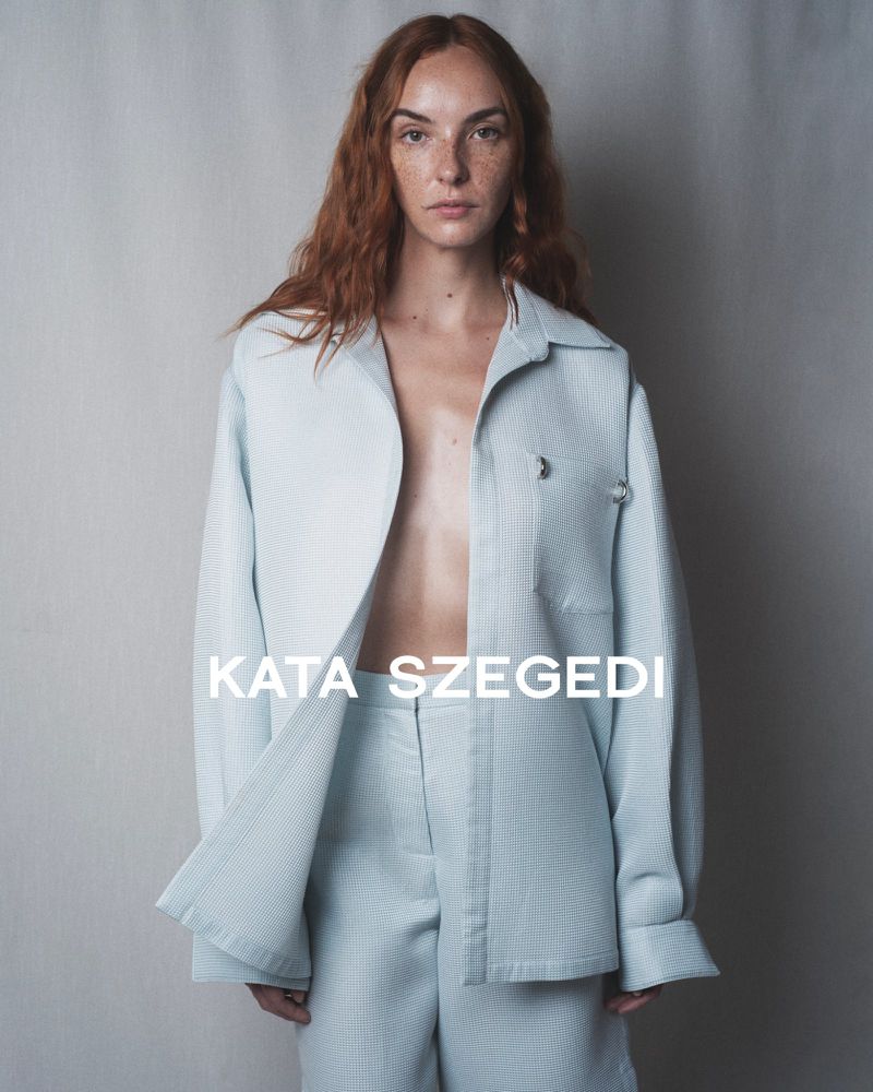 Model with long red hair wearing a light blue textured button-down shirt and matching trousers against a grey backdrop.