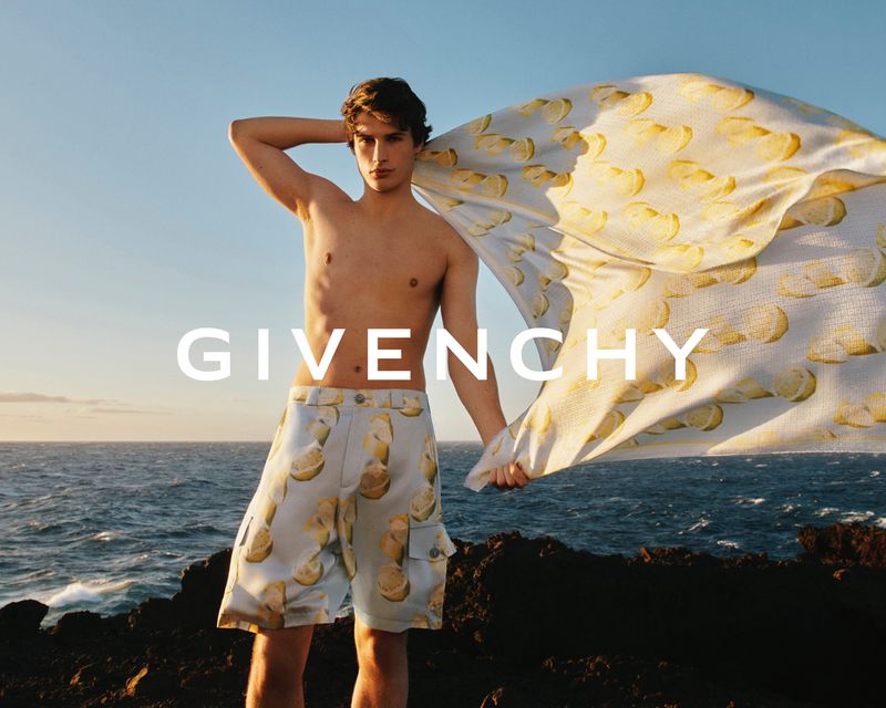 Fashion shot of a fit adult male model on a rocky coast holding a printed fabric against blue skies and ocean with brand name Givenchy.