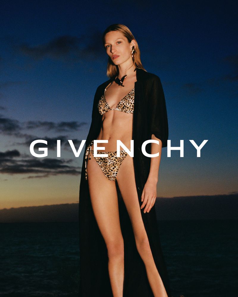 A slender model poses in a leopard-print bikini and black robe on a beach against a twilight sky in a Givenchy fashion shot.