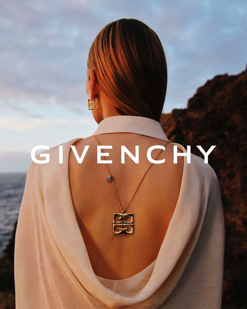 Back view of an adult woman with blonde hair, wearing a cream-colored top and gold necklace and earrings, with a coastal landscape.