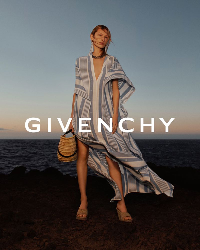 A woman models a white and blue striped dress and carries a raffia bag against a backdrop of the sea and the GIVENCHY logo.