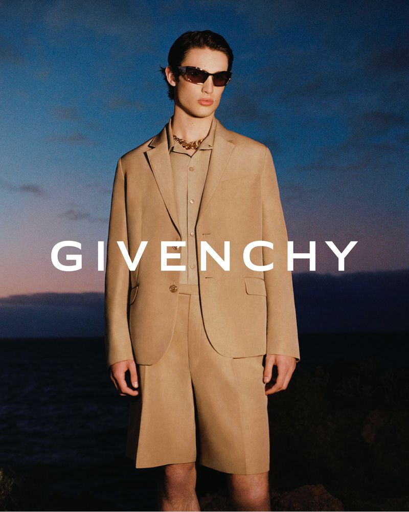 Man in tan blazer and shorts posing in front of ocean at dusk. He's wearing sunglasses and a chain necklace, with 'Givenchy' in foreground.