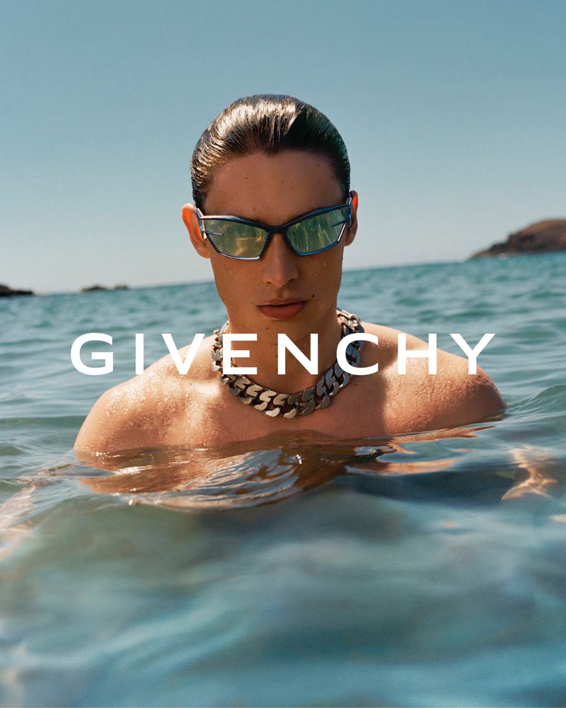 A tanned man emerges from ocean water, wearing sunglasses and a chunky necklace, with the brand GIVENCHY in white text.