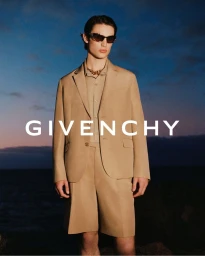 Model Unai Bartra standing in a tailored beige blazer, matching shorts, and dark sunglasses against a twilight sky for Givenchy.