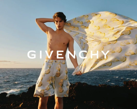 Shirtless male model wearing lemon print swim shorts and holding a printed beach towel on a rocky coast by the sea.