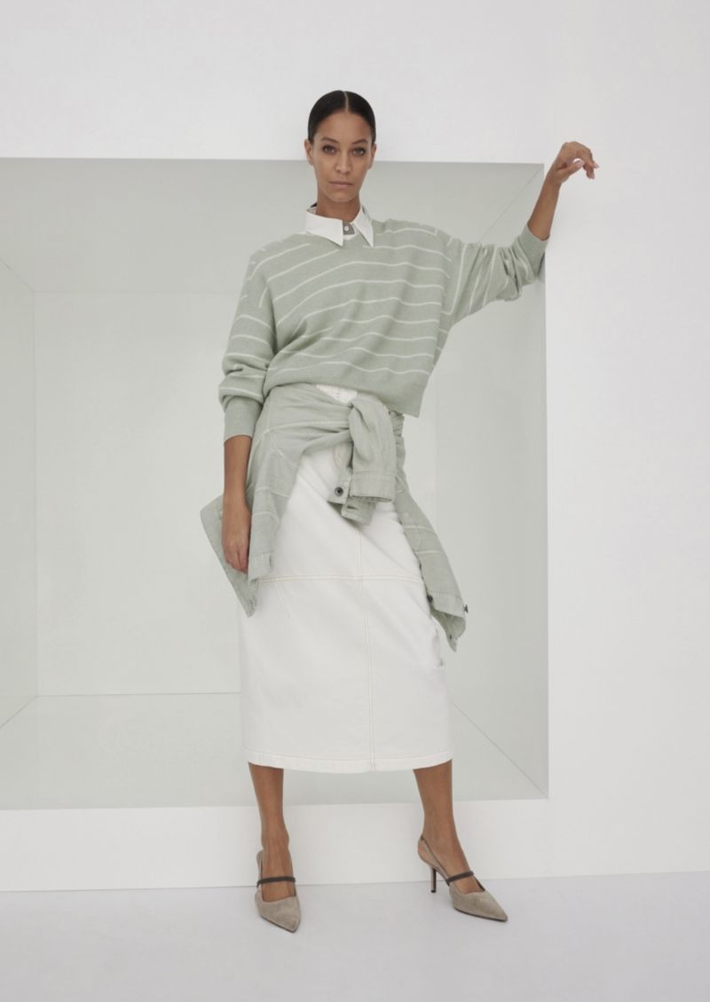 Fashion shot of a woman in a striped sweater and white skirt, leaning against a white wall, styled in minimalist tones.