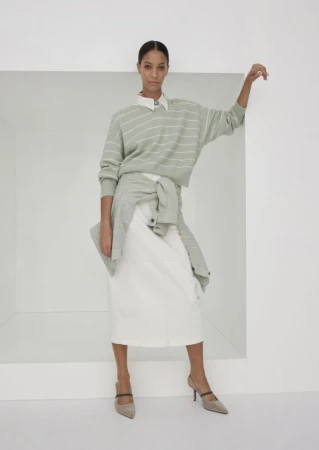 Fashion shot of a woman in a striped sweater and white skirt, leaning against a white wall, styled in minimalist tones.
