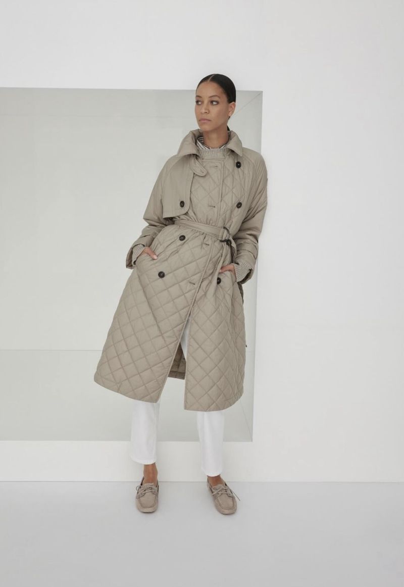 A woman poses in a quilted beige trench coat, standing against a white wall and large white square in an indoor setting.