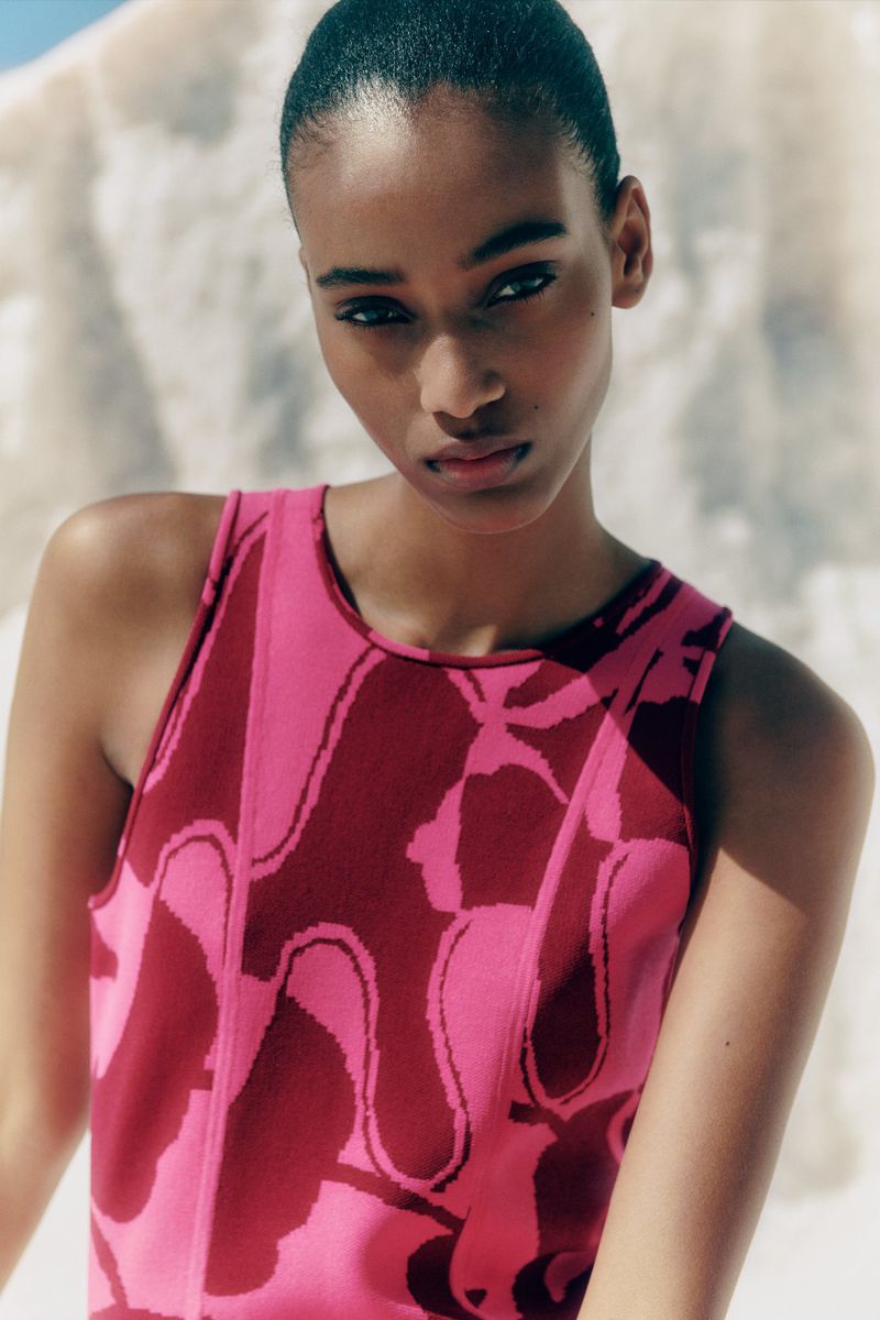 Close-up portrait of an adult woman with slicked-back dark hair wearing a pink tank top in front of a neutral-toned, textured backdrop.
