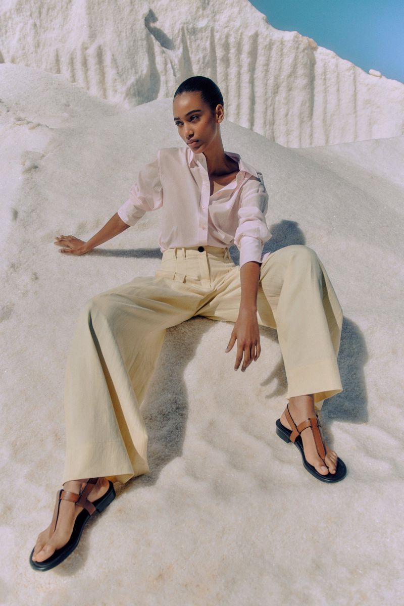 A woman in a light pink button-down and light yellow pants poses on a white surface against a clear, light blue sky.