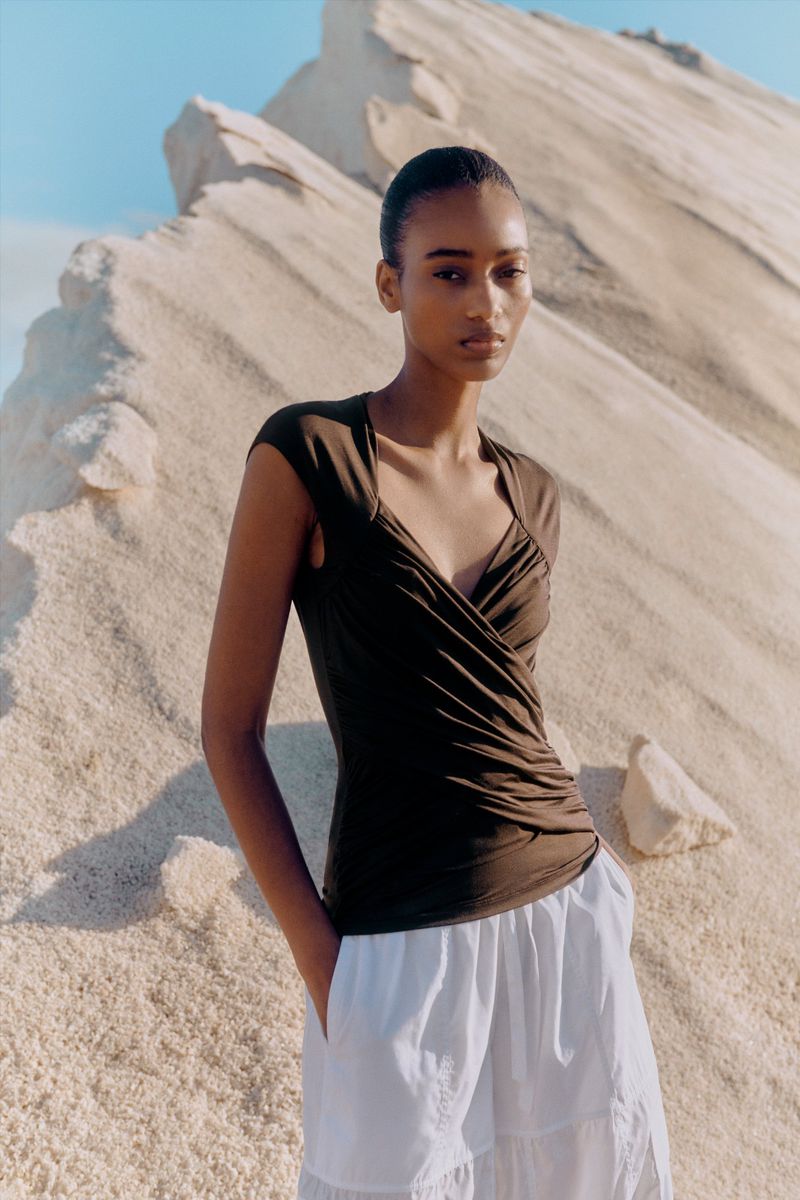 A single female model poses outdoors in a brown top and white skirt, set against a stark rock formation with a clear blue sky backdrop.