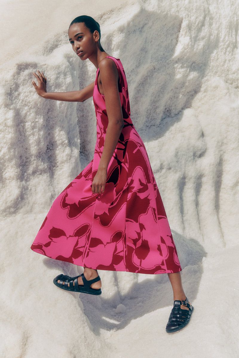 An adult woman poses in a pink and dark-red floral-patterned dress with black sandals on a white textured background, fashion shot.