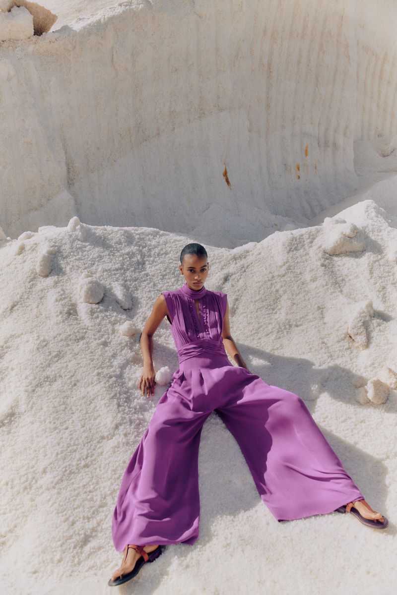 Woman in a wide-legged purple jumpsuit lying on a white, sandy beach-like surface on a bright day, fashion editorial.