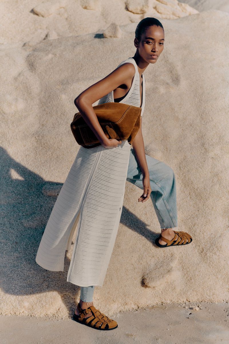 A fashionable black woman poses outdoors with a brown handbag, wearing a white knit dress, blue jeans, and brown sandals, set against a beige background.