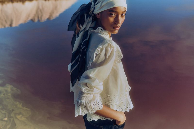 Portrait of a woman with a headscarf wearing a white blouse, standing outdoors against a blurred pink and beige background, fashion shoot.