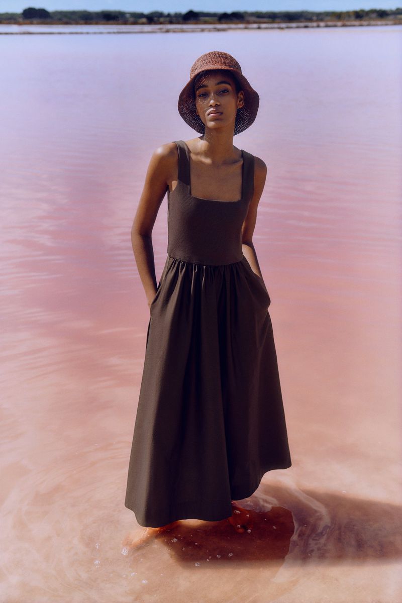 A lone female model stands in a full-length olive dress and straw hat, posing in a pink lake under natural lighting and a clear sky.