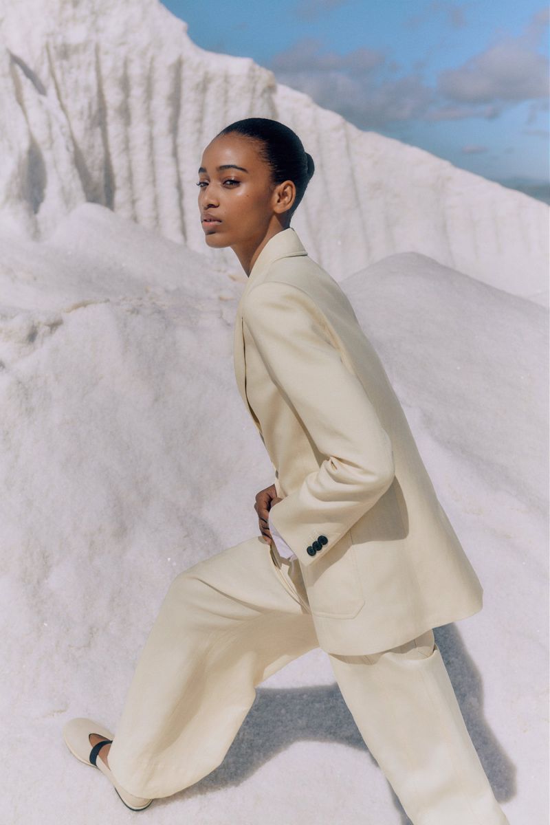 Fashionable young woman in a cream-colored suit poses in an outdoor setting with rock formations and a partly cloudy blue sky backdrop.