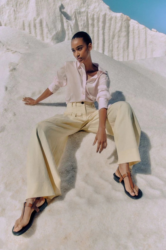Model Manuela Sanchez seated on white dunes wearing a pale pink button-up shirt, wide-leg yellow trousers, and leather sandals.
