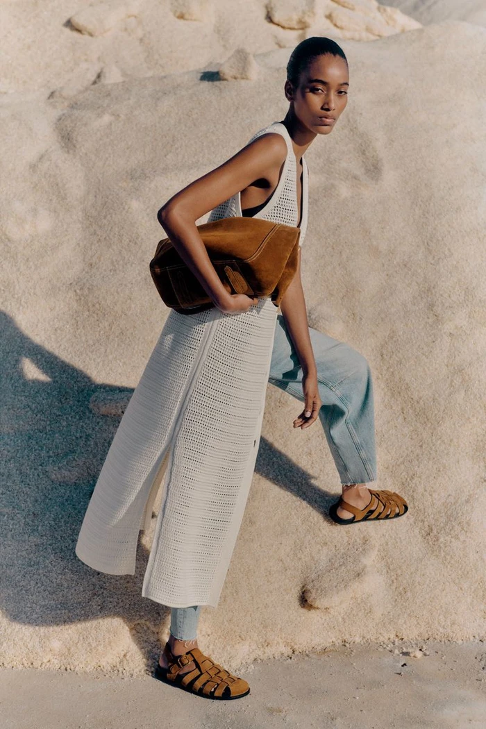 Model in cream knit tunic and blue jeans holding a brown suede bag against sand dunes