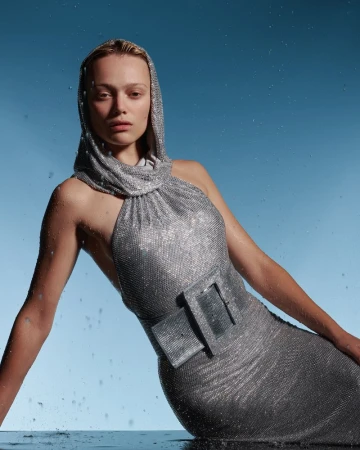 A young woman with fair skin poses in a hooded halter dress in a studio with water splashes against a blue gradient backdrop.