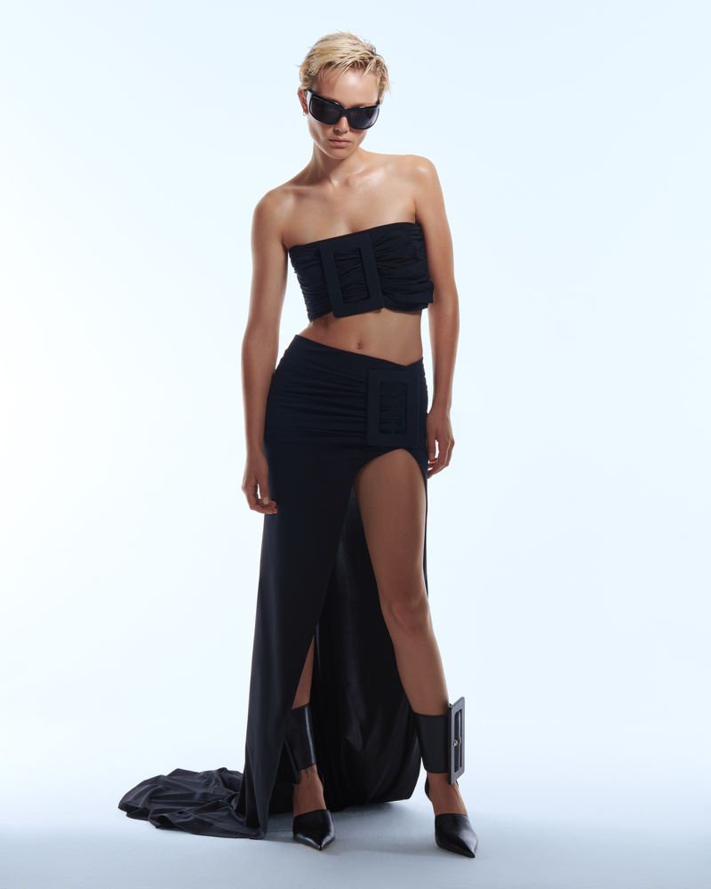 A woman in black sunglasses, a bandeau top, and a skirt with a thigh-high split poses in a studio with a white backdrop. Fashion shot.