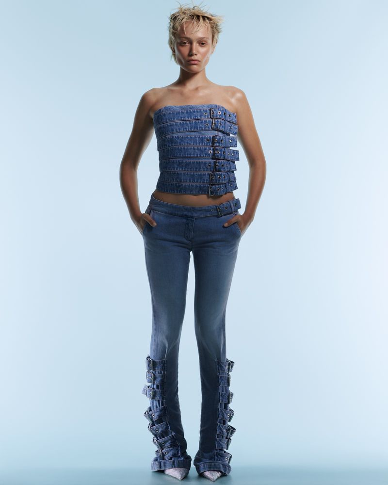 A young adult female model poses in a denim tube top and jeans with buckle details against a gradient light blue background in a studio shot.