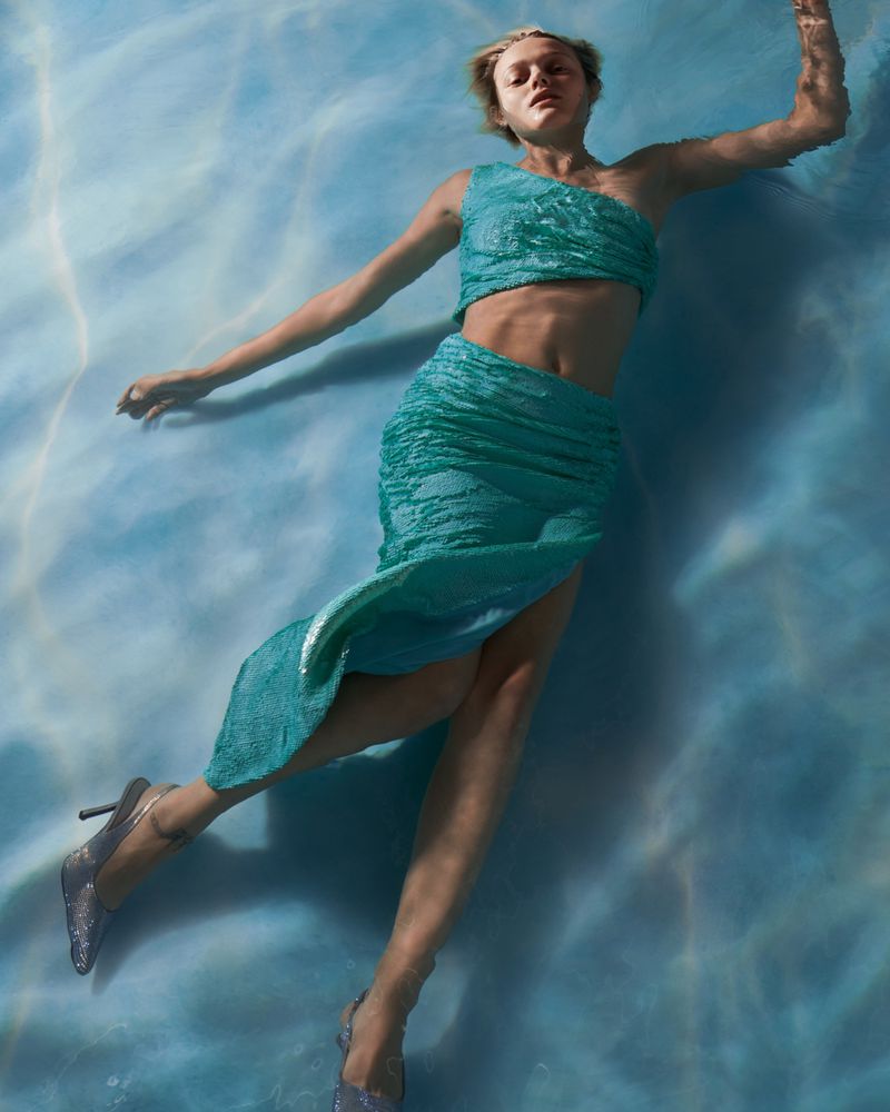 Woman in a teal, textured gown and silver high heels floats in light blue water with soft lighting highlighting her features and the water's movement.