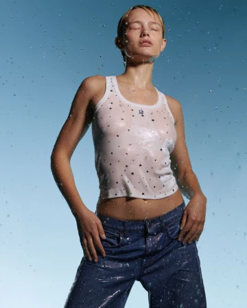 Young woman with wet hair and skin wearing a white tank top and jeans stands against a light blue background with water droplets.