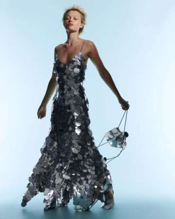 Model Simona Kust in a metallic silver disc paillette slip dress holding a matching sequined bag against a light blue studio backdrop.