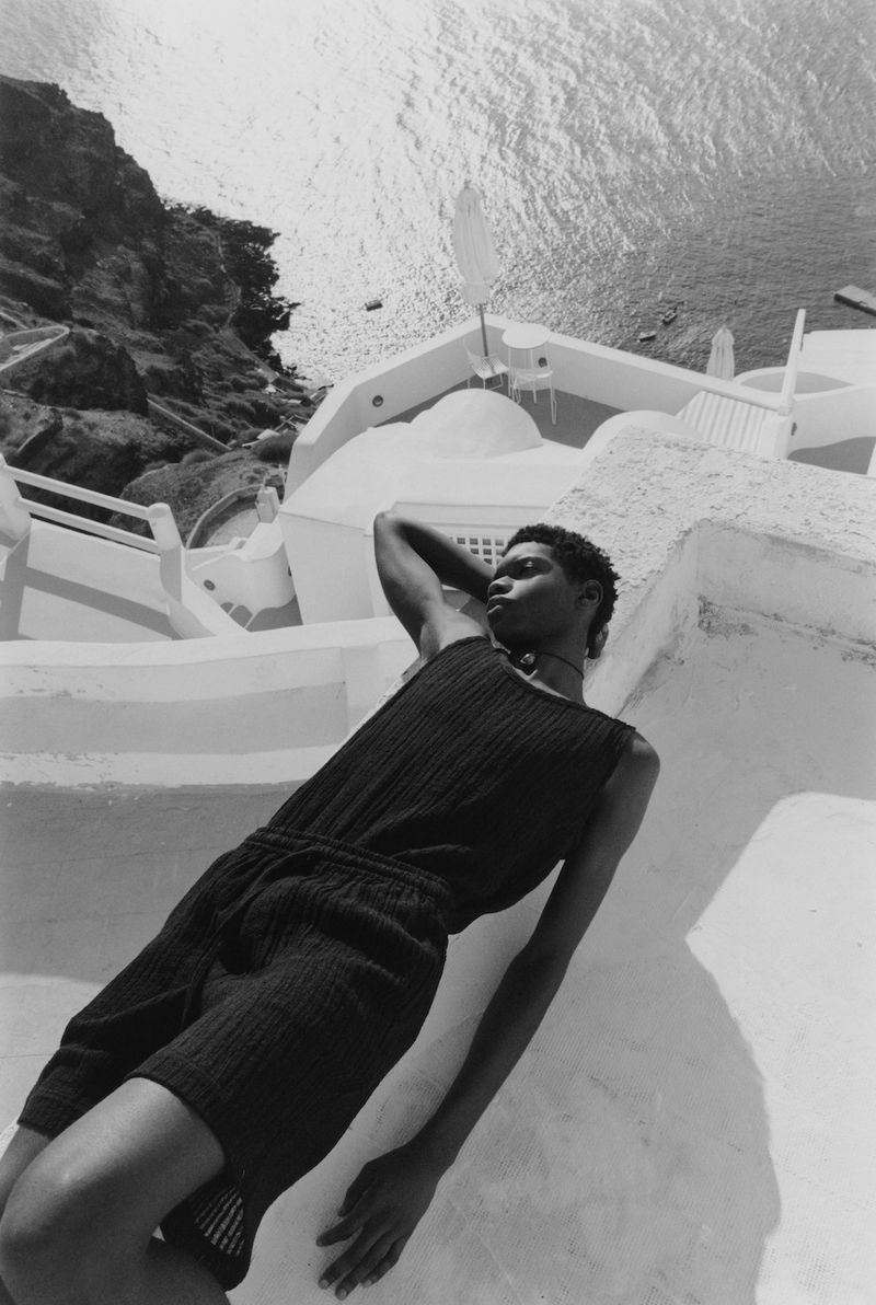 Monochromatic portrait of a young adult with dark hair in black knitted apparel lying on a white rooftop, overlooking the ocean and cliffs.