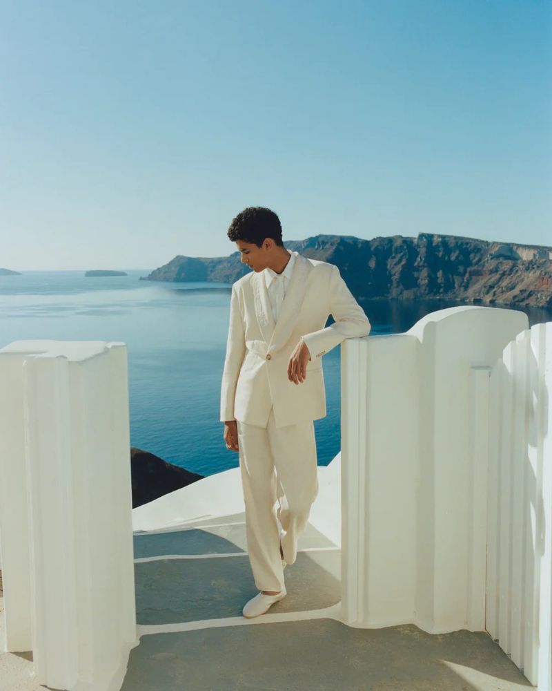 Person in an off-white suit poses on a balcony with a scenic ocean view under a clear sky in daylight for a fashion editorial.