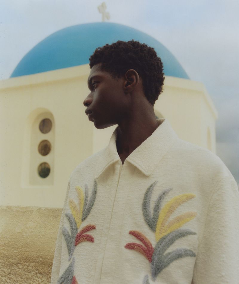 Portrait of a Black man in a patterned jacket against a white building with a blue dome, showcasing fashion and architectural details.