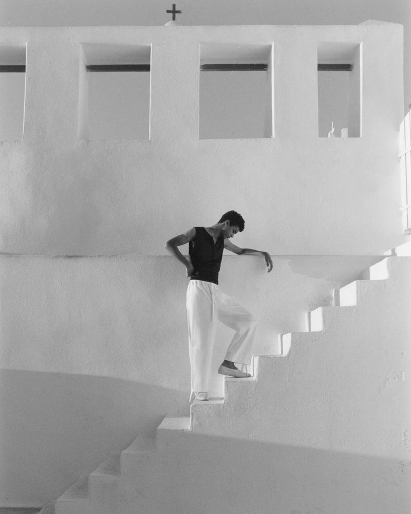 Man on white steps of building wearing dark top and white pants in black and white, architecture, cross on top of building.