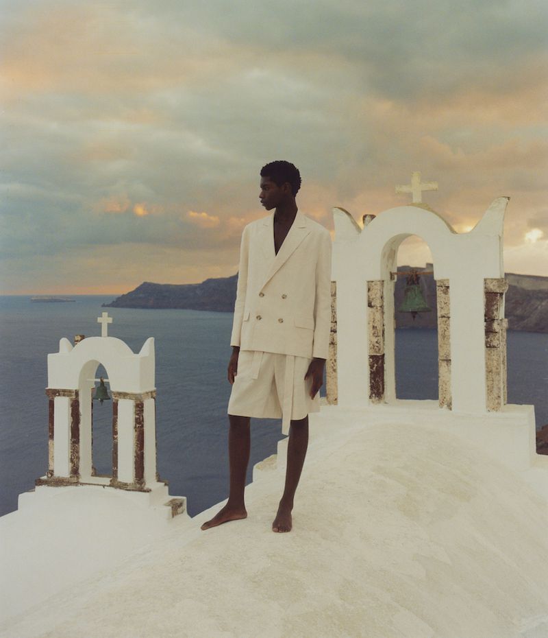 A dark-skinned man in a light-colored suit stands on a white rooftop near a small, white church against a cloudy sky and blue ocean.