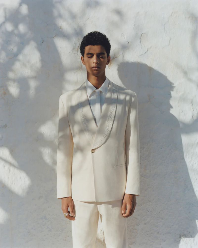 A young adult male model poses in a light-colored suit outdoors against a bright white wall with leafy shadows in a fashion shot.