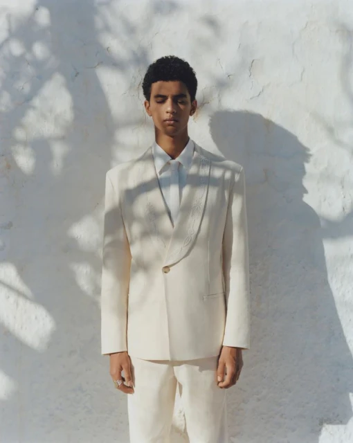 A young adult male model poses in a light-colored suit outdoors against a bright white wall with leafy shadows in a fashion shot.