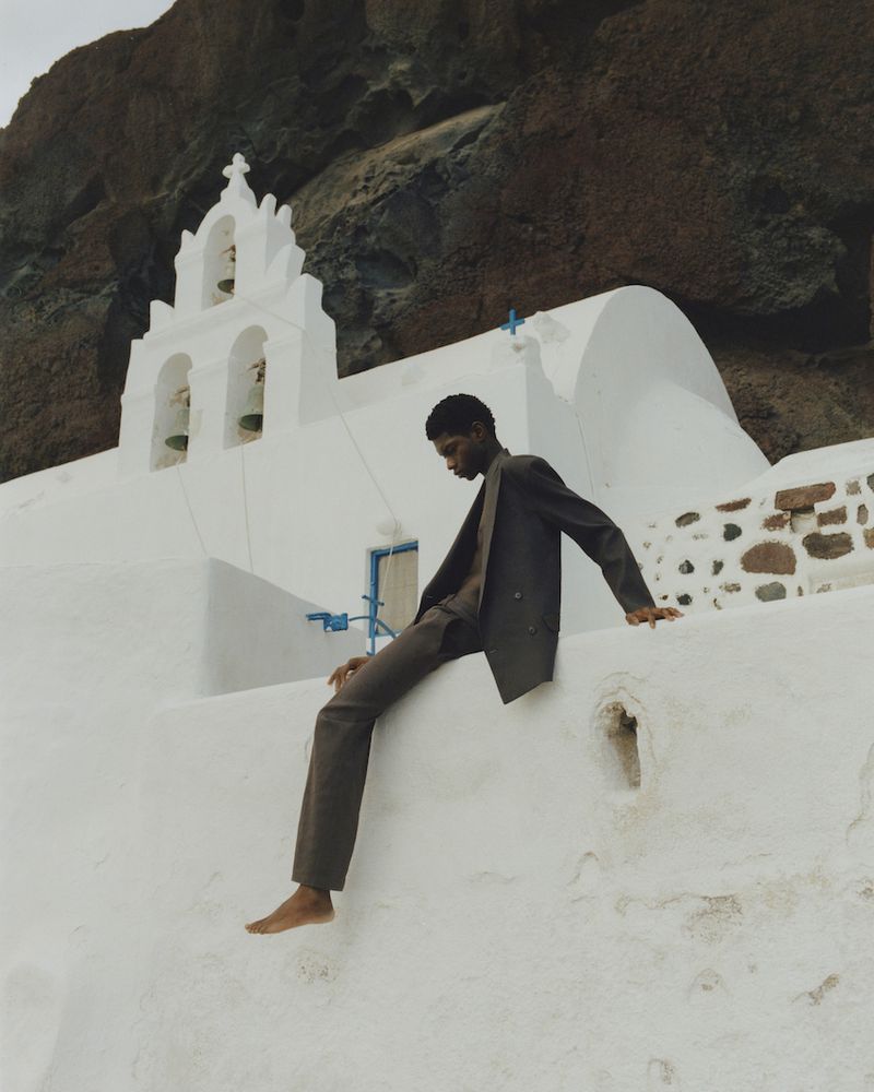 Barefoot adult man in suit sitting on wall outside white building with a mountain background. Fashion editorial photography.