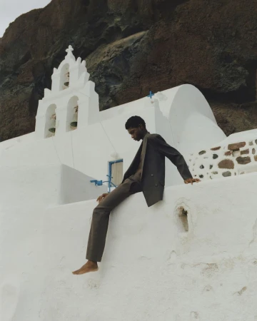 Barefoot adult man in suit sitting on wall outside white building with a mountain background. Fashion editorial photography.