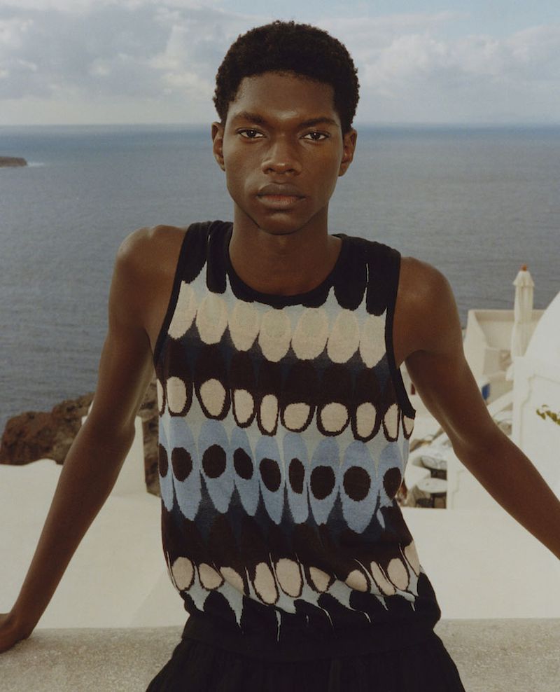 A Black male model poses in a patterned tank top against a coastal backdrop with white buildings and blue sea on a sunny day.