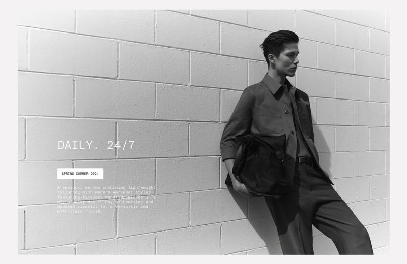 A man leans against a textured white brick wall, modeling a modern collared shirt, pants, and carrying a dark-colored casual bag.