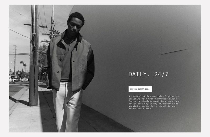 Black and white outdoor fashion shot of a Black man in a jacket and trousers, standing on a city sidewalk next to a gray wall.