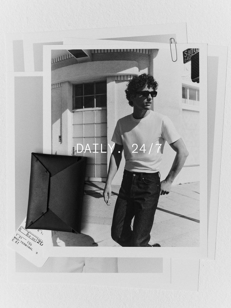 Black and white image of a man in sunglasses, jeans and a t-shirt standing in front of a building with a leather wallet.