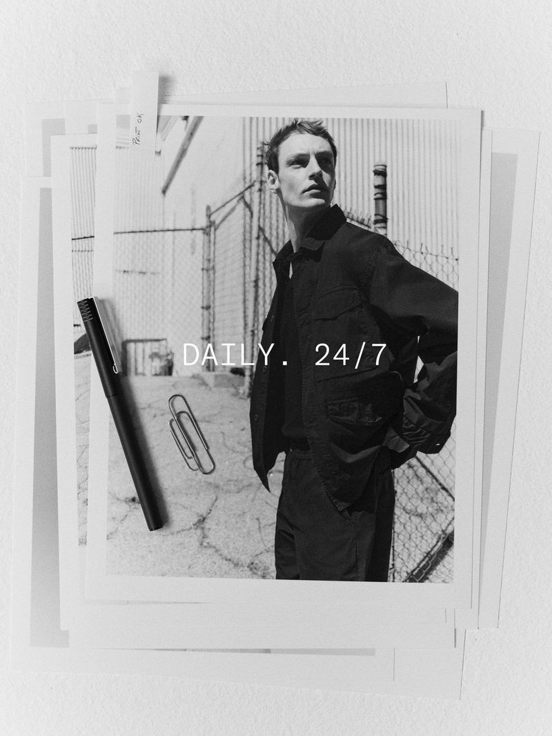 Black and white fashion photograph of a male model in a jacket, stacked on a white surface, with a pen and paper clip.