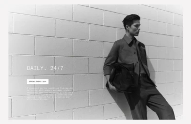 Monochrome fashion portrait of a male model leaning against a white brick wall holding a dark bag for Zara.
