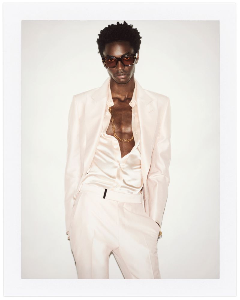 Portrait of a Black male model wearing a light pink suit, partially unbuttoned shirt, gold chain and tinted sunglasses, against a white background.