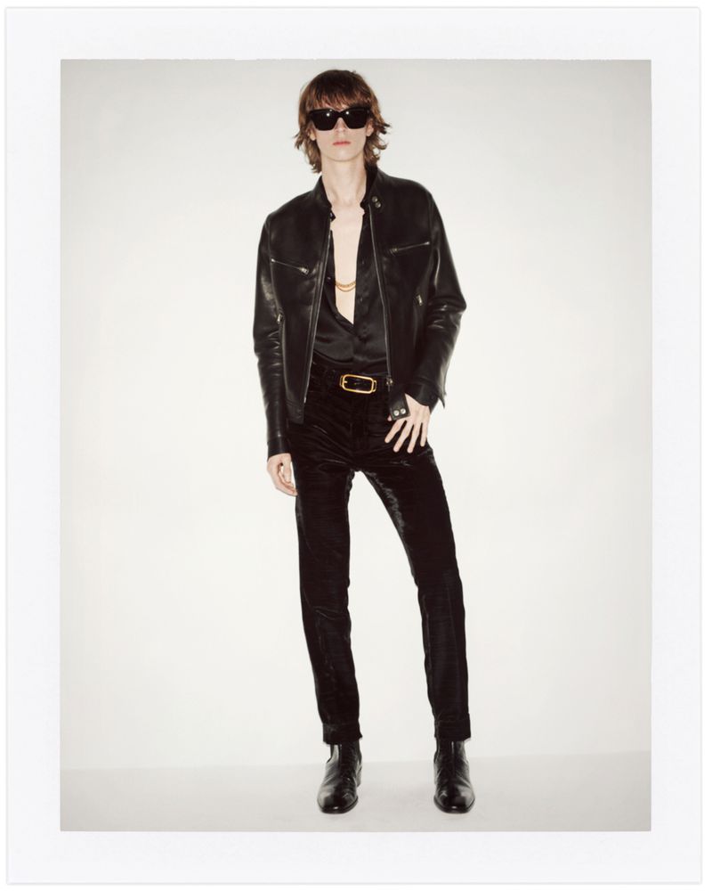 Man in a black leather jacket, black velvet pants, and sunglasses stands in a studio with a white backdrop in a full-length portrait.