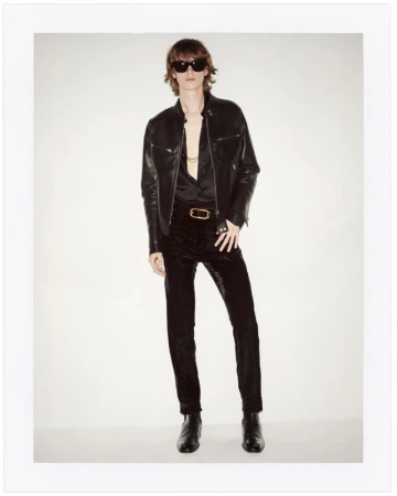 Man in a black leather jacket, black velvet pants, and sunglasses stands in a studio with a white backdrop in a full-length portrait.